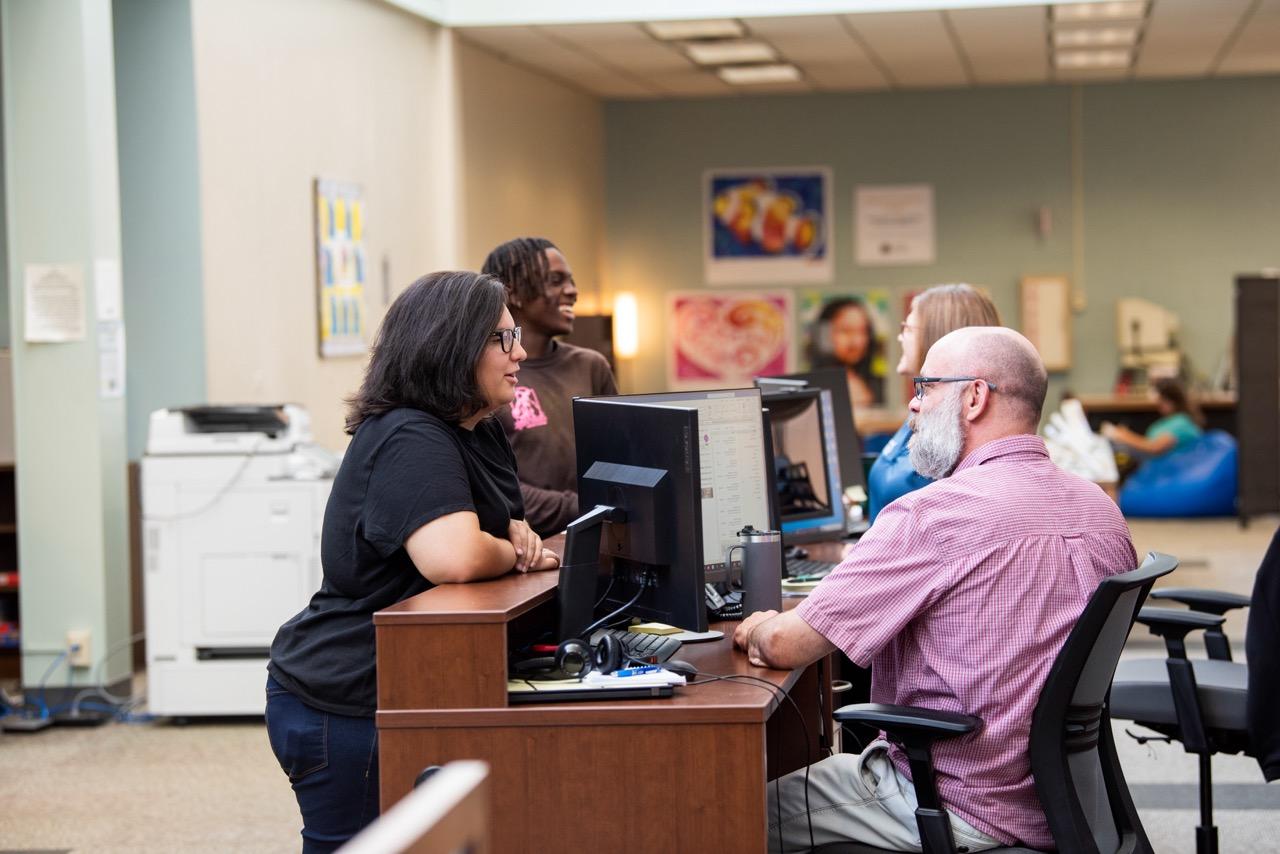 People at a desk in with computers, assisting a student with technology at the help desk.